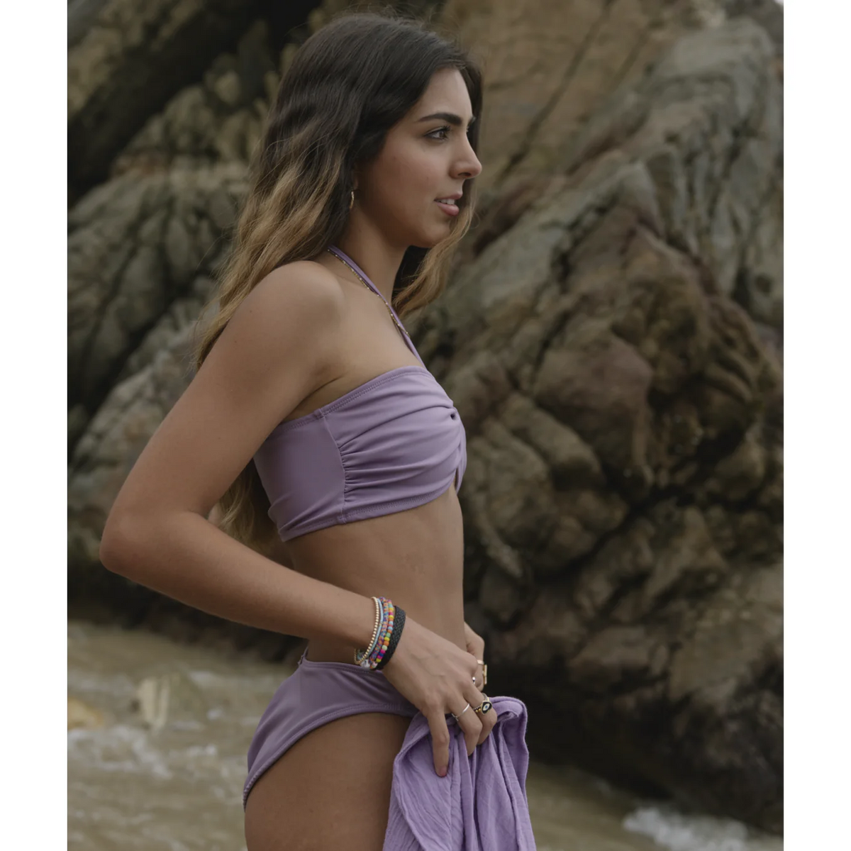 a model smiling showing how to style a jasmine bikini top on a beach with a rocky mountain in the background