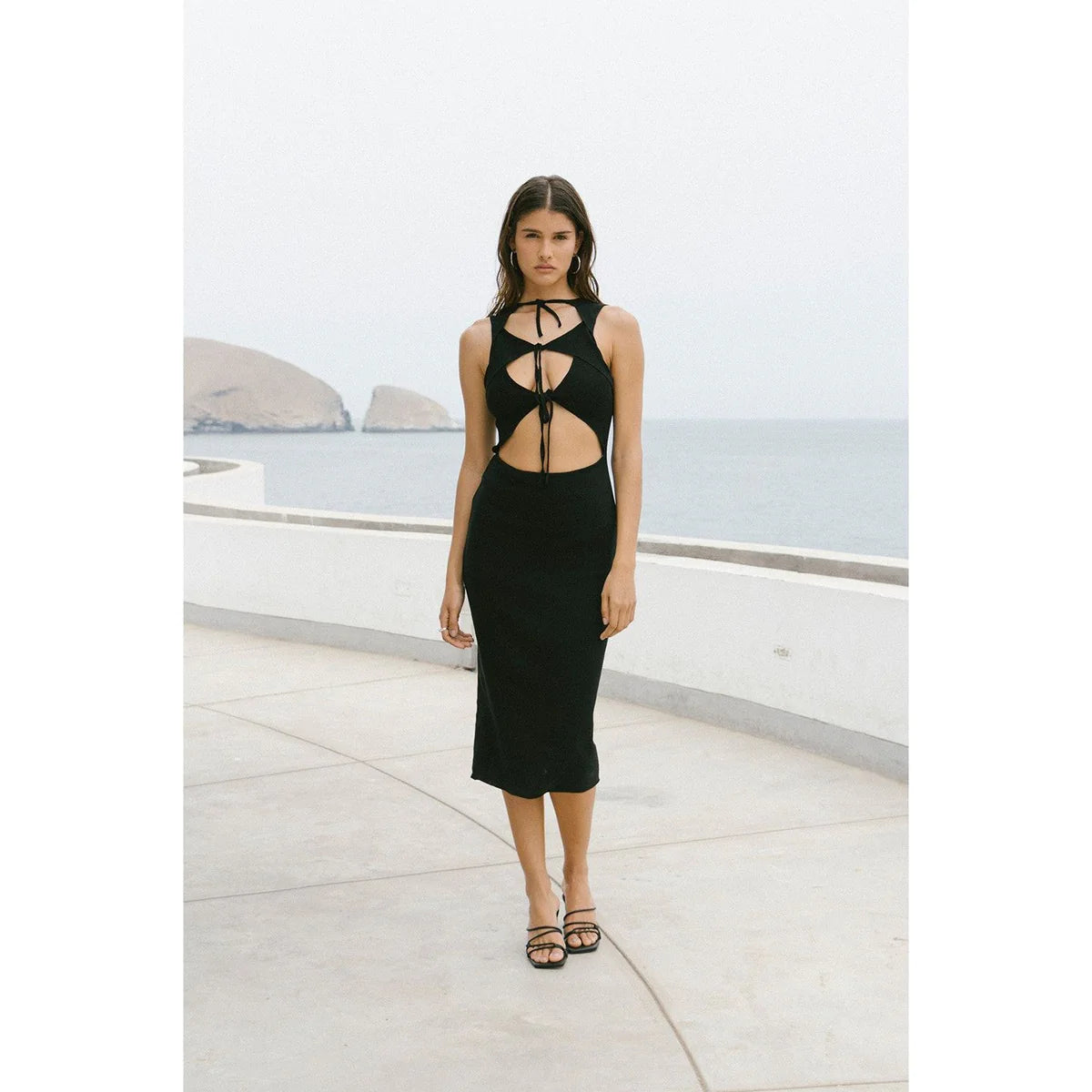 a model wearing a modern black dress with black sandals by OnPost in a pier with the sea behind